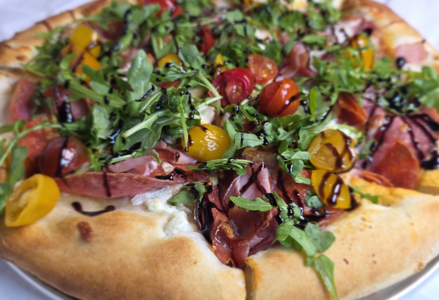 A Pizza with meat, tomatoes, arugula and a balsamic drizzel