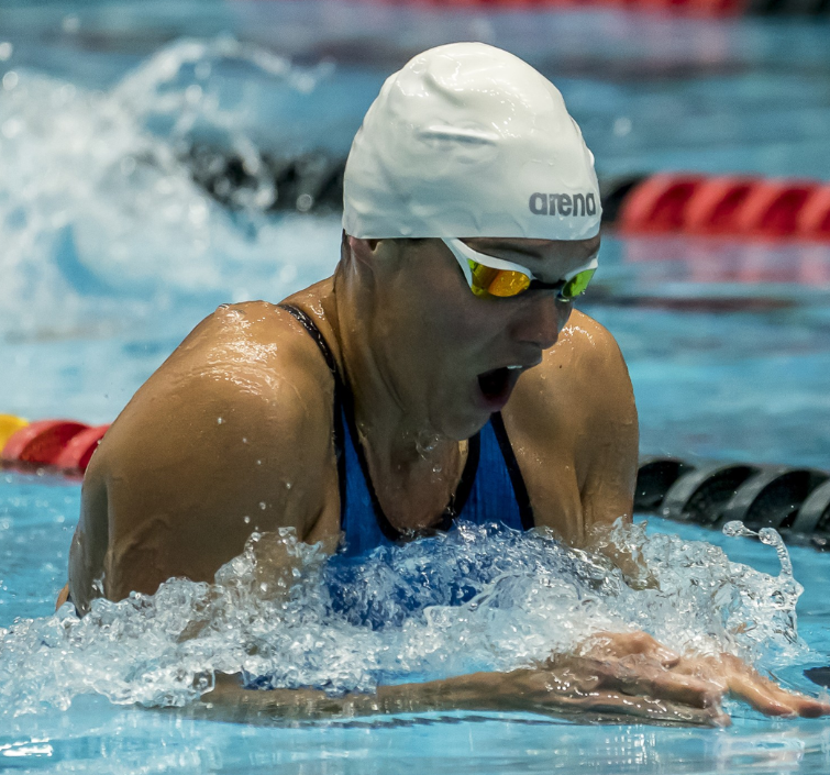 A swimmer in a white cap and goggles competes in a race, performing the breaststroke in a pool with lane dividers visible.