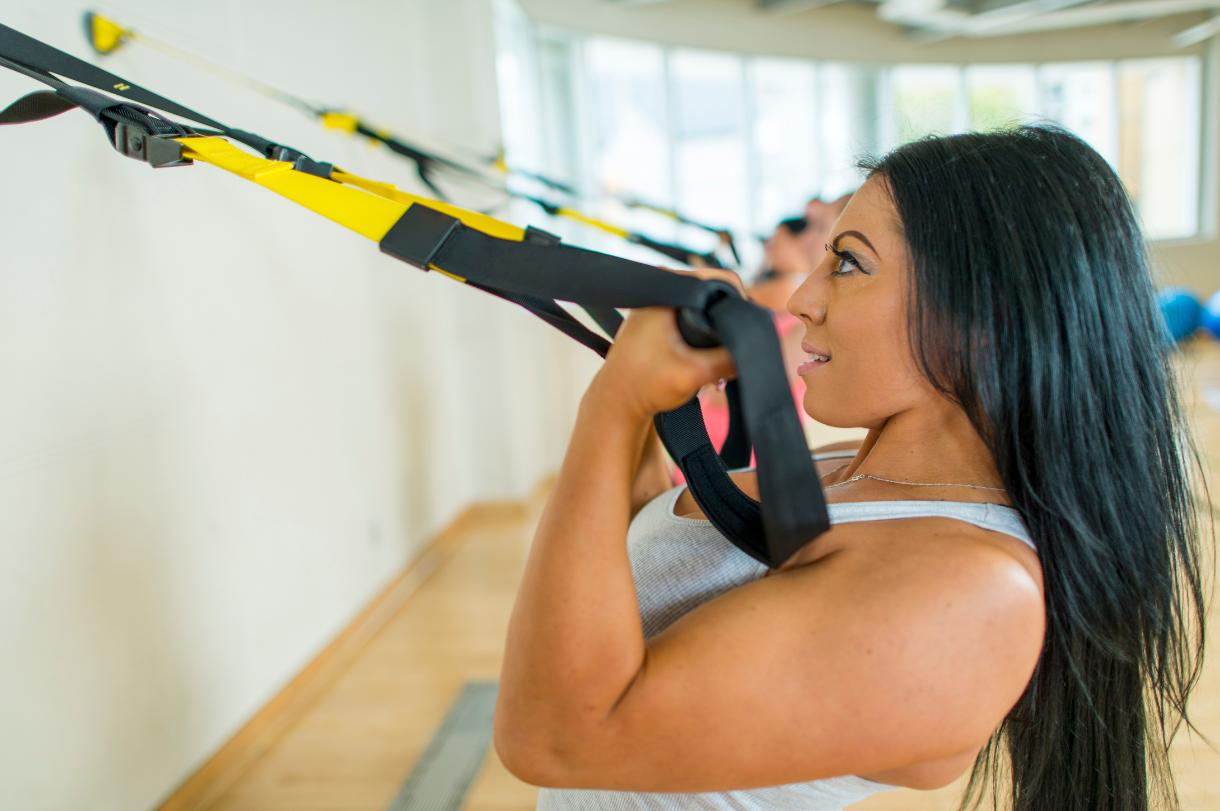 Woman exercising with suspension straps in a gym, pulling herself up while facing forward. Others are blurred in the background.