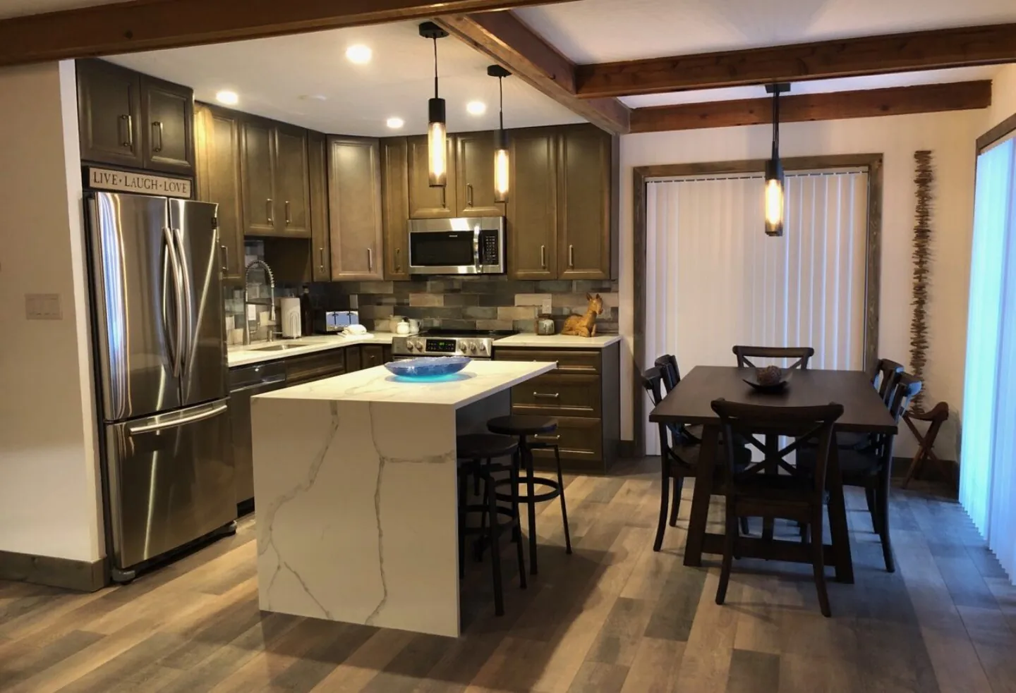 Modern kitchen with stainless steel appliances, island with a bowl, pendant lights, and adjacent dining table with six chairs. Wooden floors and beams.