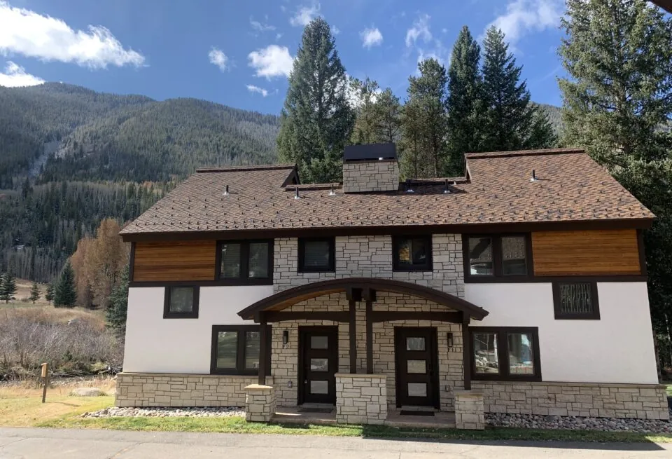 A two-story house with stone and wood accents sits in front of forested mountains under a blue sky with a few clouds.