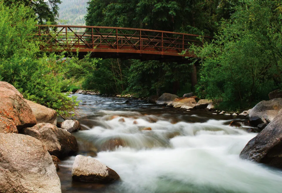 A wooden bridge spans a flowing, rocky river surrounded by lush green trees near a scenic resort in Vail.