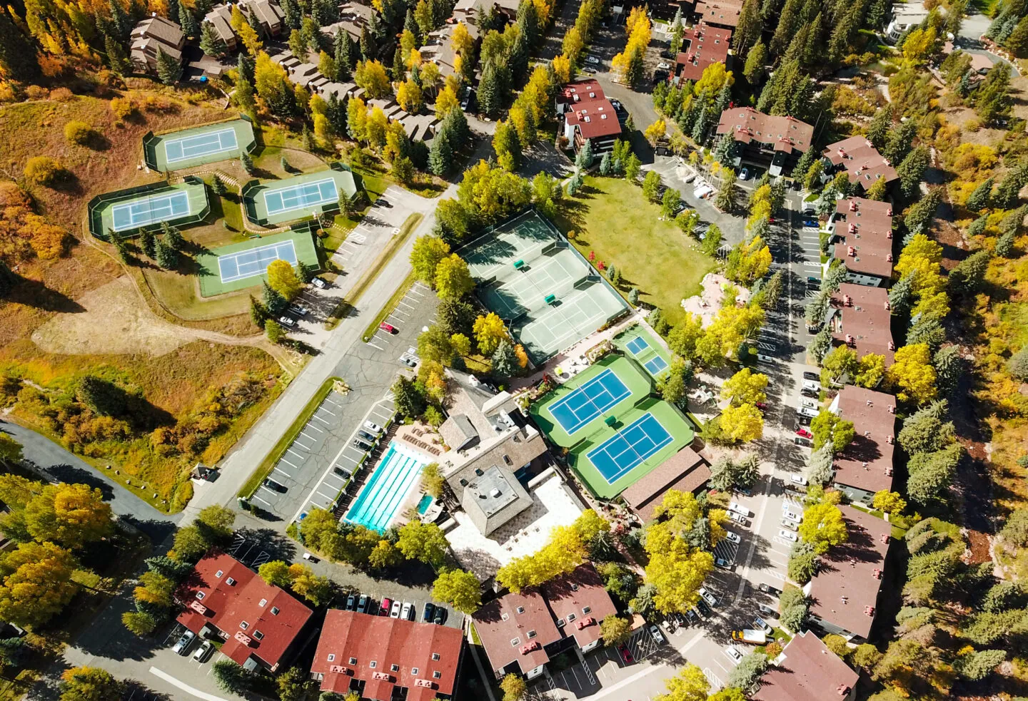 Aerial view of a tennis complex with multiple courts, a pool, and surrounding buildings among trees with autumn foliage.