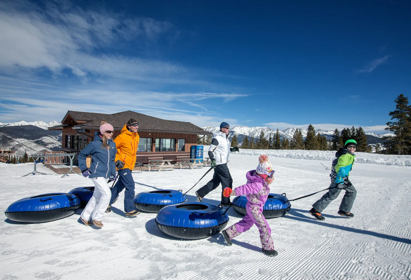 Five people, including children, pull blue snow tubes across a snowy slope with mountains and a lodge in the background under a clear blue sky.